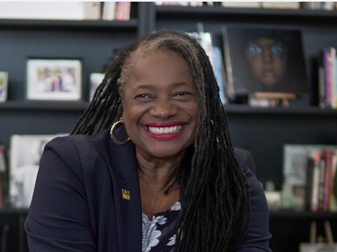 A person with long locs seated at a desk, hands clasped, wearing a navy blazer over a patterned blouse, with bookshelves and framed photos visible in the office background